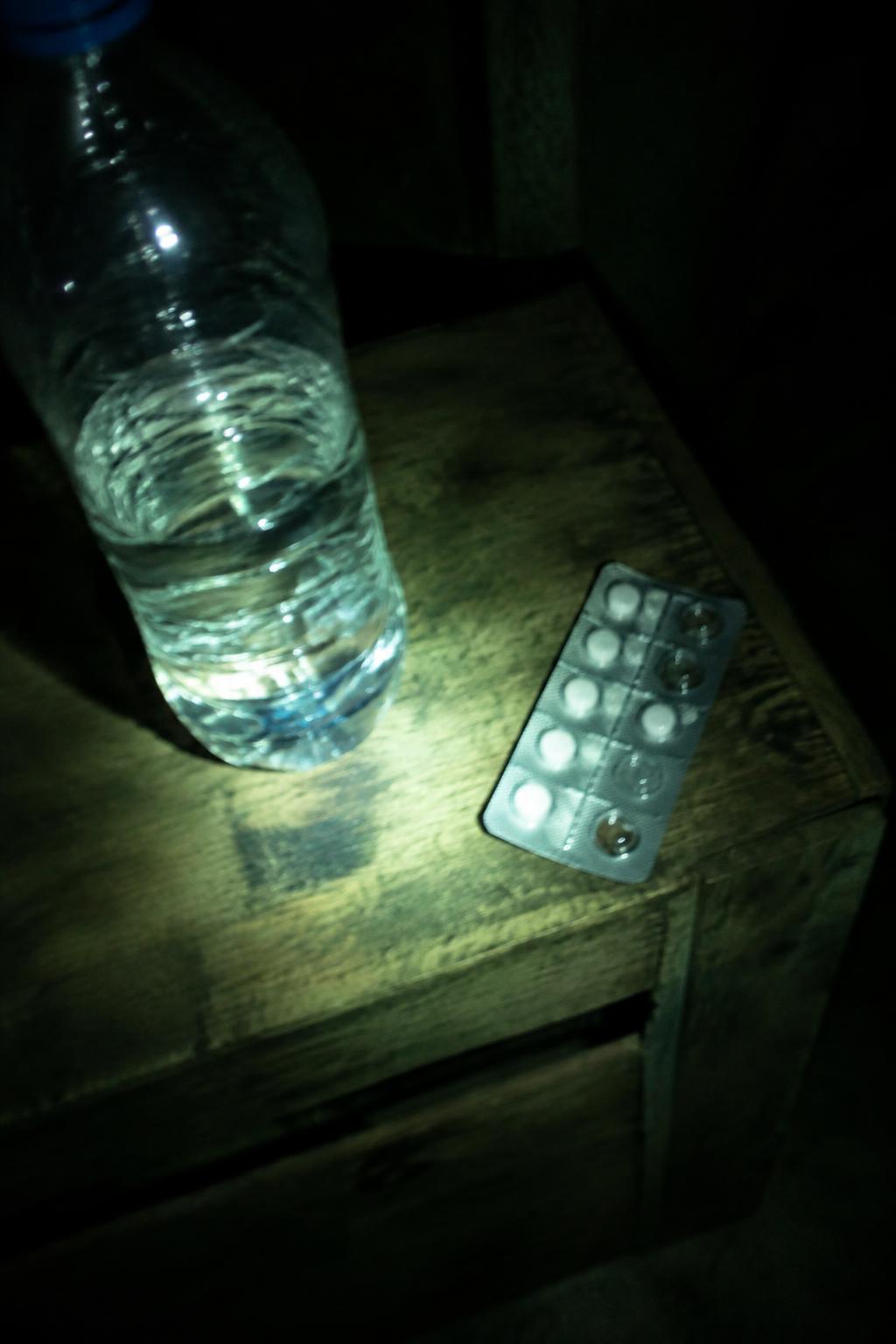 Eine halb leere Wasserflasche und eine Tablettenpackung auf einer Holzkiste, im Licht einer Taschenlampe.